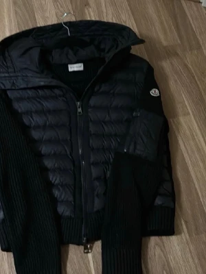 Svart pufferjacka från Moncler - Säljer en svart pufferjacka från Moncler med stickade ärmar och huva. Jackan har quiltad front, dragkedja och Moncler-logga på ärmen. Perfekt för kalla dagar och riktigt snygg till streetwear-stilen. Kan diskutera priset