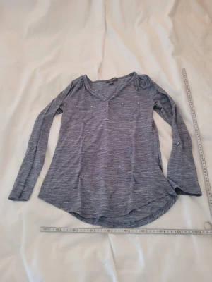 Blå långärmad topp från Primark - Mysig blåmelerad långärmad topp från Primark med v-ringning och knappdetaljer framtill. Toppen har två bröstfickor och är tillverkad i ett mjukt, tunt material som känns skönt mot huden. Perfekt till jeans eller kjol för en avslappnad look.