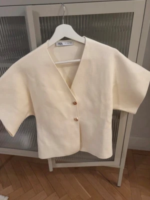 Krämvit kortärmad kofta från Zara - Stilren krämvit kofta från Zara med korta, vida ärmar och två dekorativa guldknappar framtill. Koftan har en rak passform och V-ringning, vilket ger en modern och clean look. Perfekt att slänga över en topp för en enkel men snygg outfit.