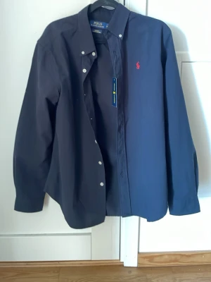 Mörkblå skjorta från Polo Ralph Lauren - Klassisk mörkblå skjorta från Polo Ralph Lauren med röd broderad logga på bröstet. Skjortan har knappar framtill och lång ärm. Tillverkad i bomull för en bekväm känsla och snygg passform.