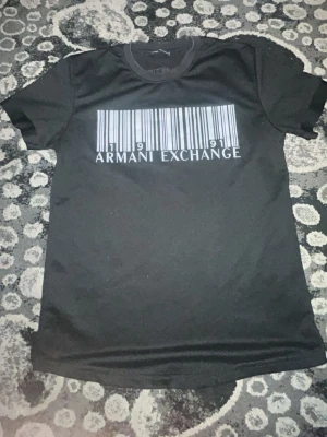 Svart Armani Exchange t-shirt - Snygg svart t-shirt från Armani Exchange med ett stort tryck av en streckkod och texten 'Armani Exchange' på bröstet. Klassisk rund hals och korta ärmar. Perfekt för dig som gillar stilrena och moderna plagg.
