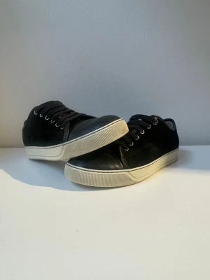 Lanvins  - Stilrena svarta sneakers med lågt skaft och vit sula. Skorna har svarta skosnören, metallfärgade öljetter och en klassisk siluett. Ovandelen verkar vara i mocka eller nubuck och sulan är i gummi. Perfekta för en clean och enkel look. De har ingen glans där fram. Priset kan diskuteras. 