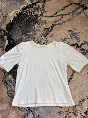 Vit topp med puffärm i bomull - Snygg vit topp med puffärm och rund halsringning. Tillverkad i mjuk bomull som känns skön mot huden. Ärmarna är halvlånga och har lätt puffad effekt vid axlarna för en trendig look. Anledningen till att jag säljer den är på grund av att toppen är inte min stil längre. 