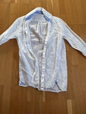 Vit linneskjorta med blå detalj - Säljer en vit skjorta i linne med klassisk krage och blå detalj på insidan av kragen. Skjortan har långa ärmar och knappar framtill. Perfekt för en avslappnad och stilren look. Dock ett litet hål på skjortan (knappt synbart).Storlek S men passar bra på M.