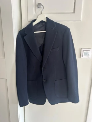 Mörkblå kavaj från John Henric - Stilren mörkblå kavaj från John Henric med två knappar framtill, klassiska slag och tre fickor. Kavajen har en diskret struktur i tyget och helfodrad insida för extra komfort. Perfekt för en dressad look.