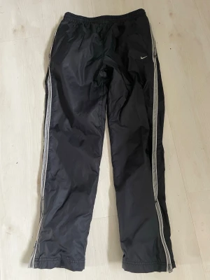 Svarta Nike mjukisbyxor med vita ränder - Svarta mjukisbyxor från Nike med vita ränder längs sidorna och liten Nike-logga på ena benet. Byxorna har resår i midjan och dragkedjor vid bensluten. Perfekta för en sportig och avslappnad stil.Barnstorlek 146-152