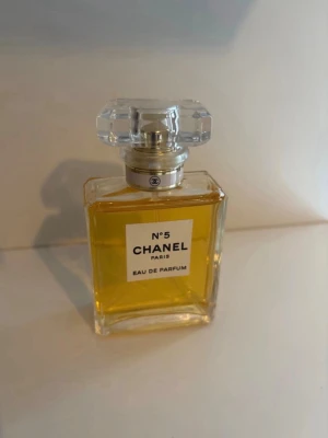 Chanel N°5 Eau de Parfum buda! - Klassisk Chanel N°5 Eau de Parfum i en elegant, rektangulär glasflaska med genomskinlig, facetterad kork. Parfymen har en gyllengul färg och flaskan pryds av en vit etikett med svart text. Perfekt för dig som gillar ikoniska dofter.