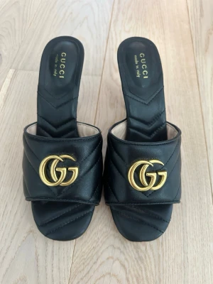 Svarta pumps med gulddetalj - Snygga svarta pumps i mjukt läder med öppen tå och bred rem över foten. Klackarna är medelhöga och har en klassisk, elegant form. Perfekta för dig som vill ha en lyxig touch till din outfit.