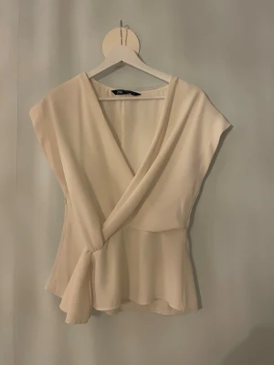 Vit omlottopp med knytning från Zara - Fin vit blus med knytning i sidan från Zara. Den är vitare i verkligheten, ljuset är lite gult. Nästan aldrig använd💕bara att skriva om du har någon fråga
