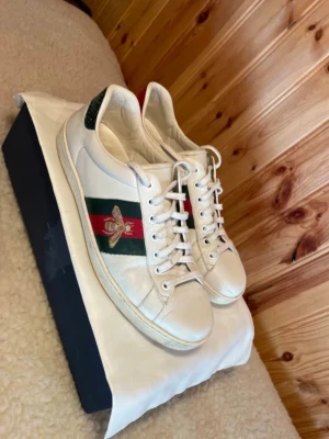 Gucci Ace sneakers uk 7/eu 41 - Stilrena vita Gucci Ace sneakers med klassiska gröna och röda ränder på sidan samt broderad guldfärgad bi. Skorna har är i uk 7/eu 41, passar 41-42. Perfekta för dig som gillar lyxiga och trendiga sneakers. Skorna är fortfarande i bra skick och har mycket kvar att ge. Väldigt bra helhetsintryck och användning/defekter syns minimalt. Hör av er vid frågor och funderingar! 