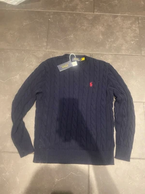 Mörkblå kabelstickad tröja Polo Ralph Lauren - Snygg mörkblå kabelstickad tröja från Polo Ralph Lauren med klassisk röd broderad logga på bröstet. Tröjan har rund halsringning, ribbade muddar och är tillverkad i mjukt stickat material. Perfekt för dig som gillar tidlös och stilren design.