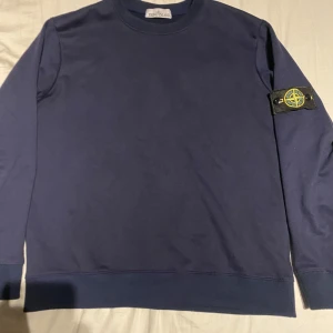 Mörkblå sweatshirt från Stone Island - Säljer en mörkblå sweatshirt från Stone Island med den klassiska logotypen på vänster ärm. Tröjan har rund halsringning, långa ärmar och ribbade muddar. Perfekt för en avslappnad och stilren look.