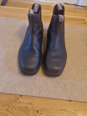 Bruna Chelsea boots från Blundstone - Säljer ett par klassiska svarta Chelsea boots från Blundstone med elastiska sidopaneler och dragflikar både fram och bak. Boots i slitstarkt läder med kontrastsömmar och robust sula. Perfekta för dig som vill ha både stil och funktion.