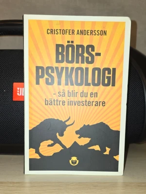 Börspsykologi av Cristofer Andersson - Boken 'Börspsykologi' av Cristofer Andersson har ett omslag i orange och svart med siluetter av en tjur och björn. Den handlar om psykologin bakom investeringar och hur du kan bli en bättre investerare genom att förstå känslor och beteenden på börsen.