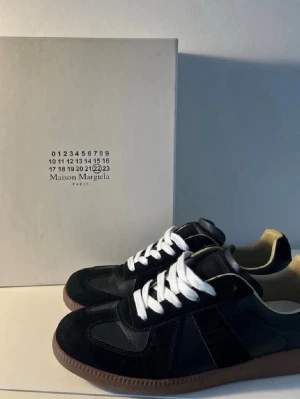 Maison Margiela svarta sneakers - Maison Margiela sneakers i svart mocka och läder med vita skosnören och klassisk gummisula i beige. Snygg och stilren design med diskreta paneler och låg silhuett. Perfekta för dig som gillar exklusiva och trendiga sneakers.