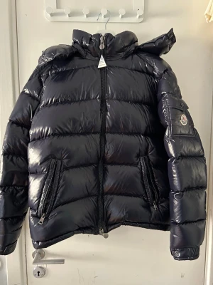 Svart dunjacka från Moncler - Snygg svart dunjacka från Moncler med glansig finish, hög krage och avtagbar huva. Jackan har två dragkedjefickor fram och en ficka med Moncler-logga på ärmen. Perfekt för kalla vinterdagar och riktigt varm tack vare dunfyllningen.