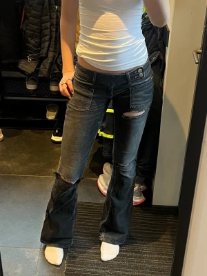 Svarta bootcut jeans med slitningar - Säljer ett par svarta bootcut jeans med slitningar på låret och knät. Jeansen har hög midja, dubbla knappar och fickor fram och bak. Snygg tvättad look och stretchigt material som sitter skönt. Perfekta för en trendig och avslappnad stil.