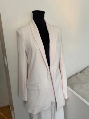 Ljusbeige kostym med kavaj och byxa - Säljer en stilren ljusbeige kostym med lång kavaj och matchande byxa. Kavajen har sjalkrage och två fickor framtill. Byxan har hög midja och ett brett knytband i midjan för extra detalj. Perfekt för dig som vill ha en modern och elegant look.