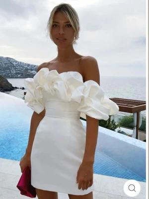 Vit sidenklänning med volanger - Trendig vit axelbandslös klänning med stora volangärmar och figurnära passform. Perfekt studentklänning! Klänningen är helt oanvänd och i perfekt skick då den inte passade mig. Den har en kort längd och markerad midja. Materialet är av satin med en elegant glans. 