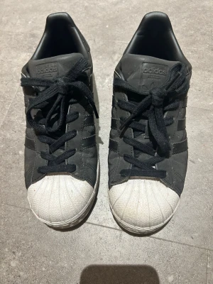 Svarta Adidas Superstar sneakers - Klassiska svarta Adidas Superstar sneakers med vita tåhättor och svarta skosnören. Skorna har de ikoniska tre ränderna på sidan och svart polyester. Sparsamt använda men väldigt bekväma. Storleken är som en vanlig 38. 
