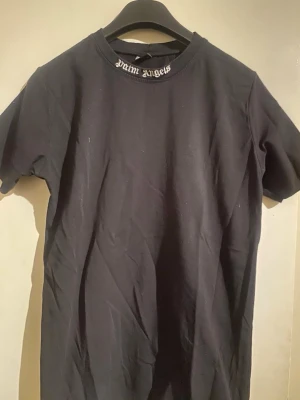 Svart Palm Angels t-shirt - Säljer en svart t-shirt från Palm Angels med logga i vit gotisk stil både på kragen och stort på ryggen. T-shirten har rund hals och korta ärmar. Materialet är mjuk bomull som känns skön mot huden. Perfekt för dig som gillar streetwear och vill sticka ut.