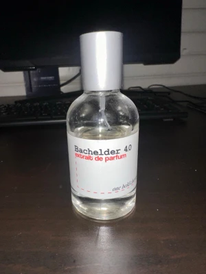 Bachelder 4.0 Extrait de Parfum - En stilren parfymflaska från Bachelder med silverfärgat lock och transparent glas. Flaskan har en enkel vit etikett med röd och grå text där det står 'Bachelder 4.0 extrait de parfum'. Perfekt för dig som gillar minimalistisk design.