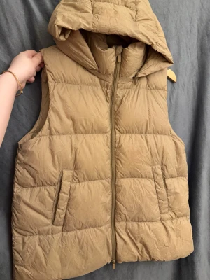 Beige dunväst med huva - Snygg beige dunväst med stor, vadderad huva och dragkedja framtill. Västen har två snedställda fickor och quiltad design med breda paneler. Perfekt för lager-på-lager och ger en avslappnad streetwear-look.