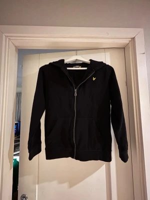 Svart zip hoodie från Lyle & Scott - Svart hoodie med dragkedja från Lyle & Scott. Klassisk modell med huva, två fickor framtill och gul broderad logga på bröstet. Tillverkad i mjuk bomullsblandning och har ribbade muddar vid ärmar och nederkant.