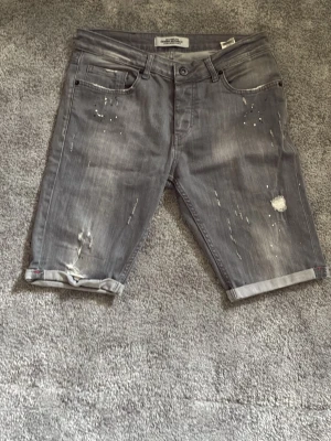 Grå jeansshorts med slitningar - Säljer ett par grå jeansshorts med slitna detaljer och uppvikta benslut. Shortsen har fem fickor och coola slitningar framtill och baktill för en avslappnad streetstil. Perfekta för dig som gillar en edgy look.