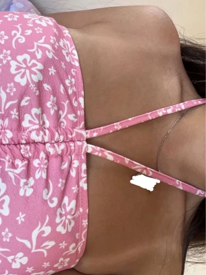 Rosa blommig halterneck topp - Rosa topp med vita blommor och smala halterneckband som knyts i nacken. Toppen har rynkdetalj framtill som går att justera och är figurnära med croppad längd. Skönt material,  perfekt för sommaren. Tror den är i storlek S/M men har klippt av lappen. Använd några gånger men inga defekter eller slitage. Kan självklart diskutera pris, hör av dig vid frågor!🩷