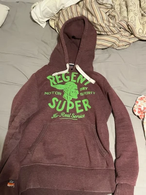Vinröd hoodie från Superdry - Säljer en vinröd hoodie från Superdry med grön text och tryck på bröstet. Hoodien har huva med vita snören och en stor magficka. Tillverkad i mjukt material som passar perfekt för en avslappnad stil.