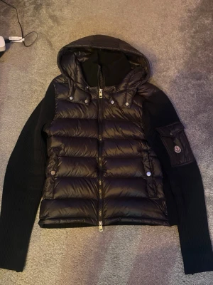 Moncler cardigan - Snygg svart dunväst från Moncler med stickade svarta ärmar och huva. Västen har dragkedja framtill, två fickor med tryckknappar och en ärmficka med Moncler-logga. Perfekt för lager-på-lager och streetstyle.