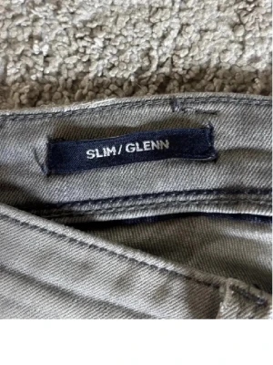 Slim Glenn grå slim fit jeans - Snygga grå slim fit jeans i modellen Slim/Glenn. Jeansen har klassisk femficksdesign, bälteshällor och dekorativa sömmar på bakfickorna. Perfekta för dig som gillar en smalare passform och stilren look.