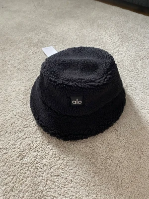 Svart teddy bucket hat från Alo - Snygg svart bucket hat från Alo i mjukt teddy-material. Hatten har en klassisk form med bred kant och en liten Alo-logga framtill. Perfekt för dig som vill ha en trendig och mysig accessoar till höst och vinter.