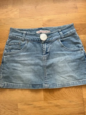 Blå jeanskjol från Madonna  - Så snygg jeanskjol från Madonna med fem fickor, bälteshällor och en stor knapp framtill. Kjolen har en kort passform och är gjord i mjuk denim med snygga sömdetaljer på bakfickorna. Perfekt till sneakers eller boots!