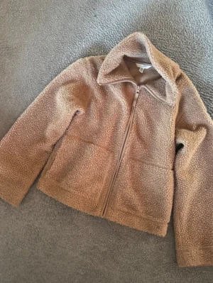 Beige teddyjacka med dragkedja - Mysig beige teddyjacka med dragkedja framtill och två stora fickor. Jackan har en klassisk krage och är gjord i mjukt, fluffigt teddy-material som håller dig varm. Perfekt för kyliga dagar och enkel att matcha med olika outfits.