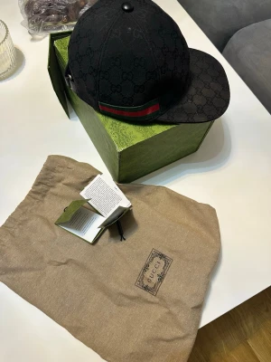 Svart Gucci keps med GG-mönster - Snygg svart keps från Gucci med klassiskt GG-monogram över hela kepsen och en grön-röd rand på sidan. Justerbar rem bak för perfekt passform. Kepsen kommer med original dustbag och box. Perfekt accessoar för dig som vill sticka ut.