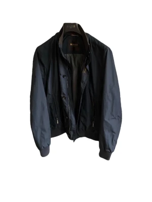 Svart jacka - Stilren svart vindjacka från Moorer med dragkedja och knappar framtill. Jackan är äkhetscheckad via Vestaire Collective. Pris: 6000kr, men kan sänkas vid snabb affär!