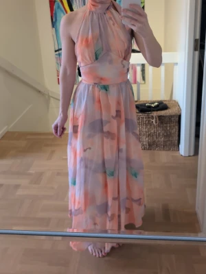Lång pastellfärgad halterneckklänning - Säljer en lång halterneckklänning i mjuka pastellfärger med abstrakt mönster. Klänningen har en hög hals, markerad midja och ett flytande, luftigt tyg som ger en elegant siluett. Perfekt för sommarens alla tillfällen!