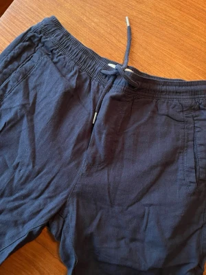 Mörkblå byxor i linne med snörning - Snygga mörkblå byxor med elastisk midja och snörning framtill. Byxorna har sidofickor och en bakficka med knapp. Perfekta för varma dagar och ger en avslappnad look.