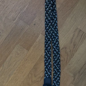  flätat bälte från John henric  - Snyggt flätat bälte med randigt mönster i svart, grått och vitt. Bältet har en metallspänne och detaljer i svart.