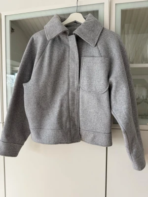 Grå ulljacka med stor ficka - Snygg grå jacka med rak passform och stor bröstficka. Jackan har bred krage, dold dragkedja och tryckknappar framtill samt fodrad insida för extra komfort. Perfekt för dig som gillar minimalistisk och stilren design.
