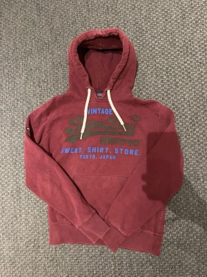 Vinröd Superdry hoodie med tryck(RESERVERAD) - Vinröd hoodie från Superdry med stor huva och vita dragsnören. Framsidan har ett stort tryck i grönt, blått och vitt med text och japanska tecken. Klassisk känguruficka framtill och mjukt bomullsmaterial som känns skönt mot huden.
