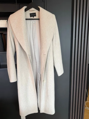 Beige ullkappa från STOCKH LM - Snygg beige kappa från STOCKH LM i storlek 36. Kappan har en oversized krage, diskreta fickor och stängs med knapp framtill. Tillverkad i mjuk ullmix med ett elegant, mönstrat innerfoder. Perfekt för kyliga dagar och ger en clean look.