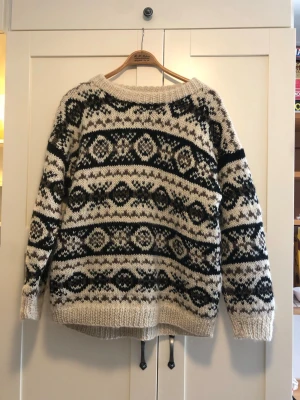 Beige och svart stickad ulltröja - Säljer en handgjord, chunky stickad tröja från Nepal i 100% ull. Tröjan är beige med svart mönster, har rund halsringning och långa ärmar. Perfekt för kalla dagar och ger en mysig, retro vibe.
