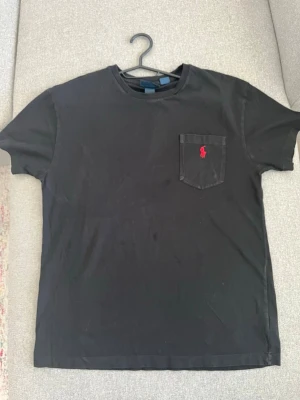 Svart t-shirt från Polo Ralph Lauren - Klassisk svart t-shirt från Polo Ralph Lauren med röd broderad logga på bröstfickan. T-shirten har rund halsringning och korta ärmar. Tillverkad i mjuk bomull för en skön känsla och enkel stil.