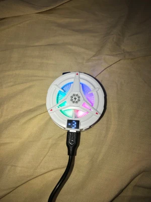 RGB USB-fläkt med LED-belysning - Säljer en rund USB-fläkt i vit plast med färgglad RGB LED-belysning. Fläkten har en futuristisk design med tre armar och en liten digital display. Perfekt för gaming-setup eller skrivbordet för extra cool effekt.