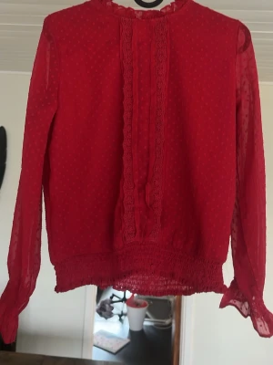Röd blus med fina detaljer  - Storlek 146/152. Trendig röd blus med fina detaljer som prickar och volanger. Du knäpper blusen med en knapp bak. Tröjan har en svart fläck på vänster ytterarm. Denna tröja passar perfekt till jultillfällen.