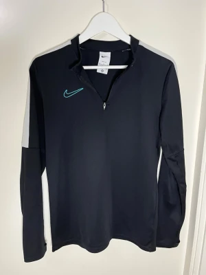 Svart Nike träningströja med dragkedja - Svart långärmad träningströja från Nike med halv dragkedja framtill och turkos logga på bröstet. Tröjan har gråa detaljer på axlarna och är tillverkad i ett stretchigt funktionsmaterial som passar perfekt till träning.