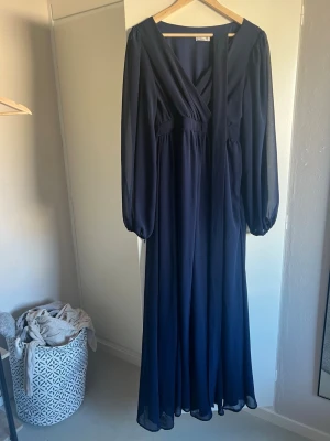 Mörkblå långklänning med v-ringning - Elegant mörkblå långklänning med v-ringad omlott framtill och långa, löst sittande ärmar. Klänningen har markerad midja och är tillverkad i ett lätt, flytande tyg som ger ett snyggt fall. Perfekt för dig som gillar stilrena och klassiska plagg.